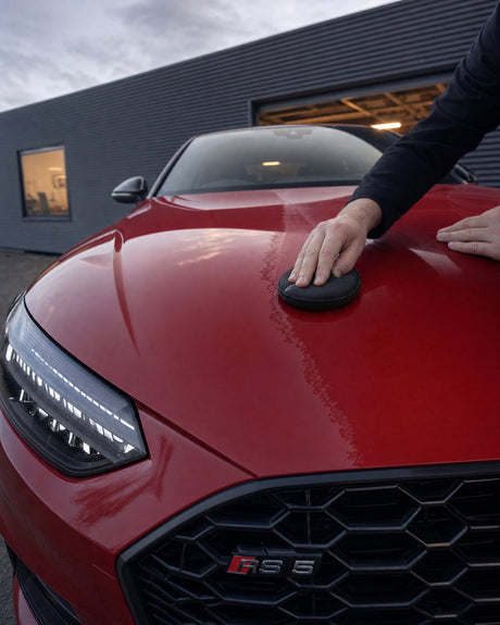 Daniel wachst mit dem Applikator die rote Haube eines Audi RS 5; eine Bahn glänzt schon tief, daneben noch matt.